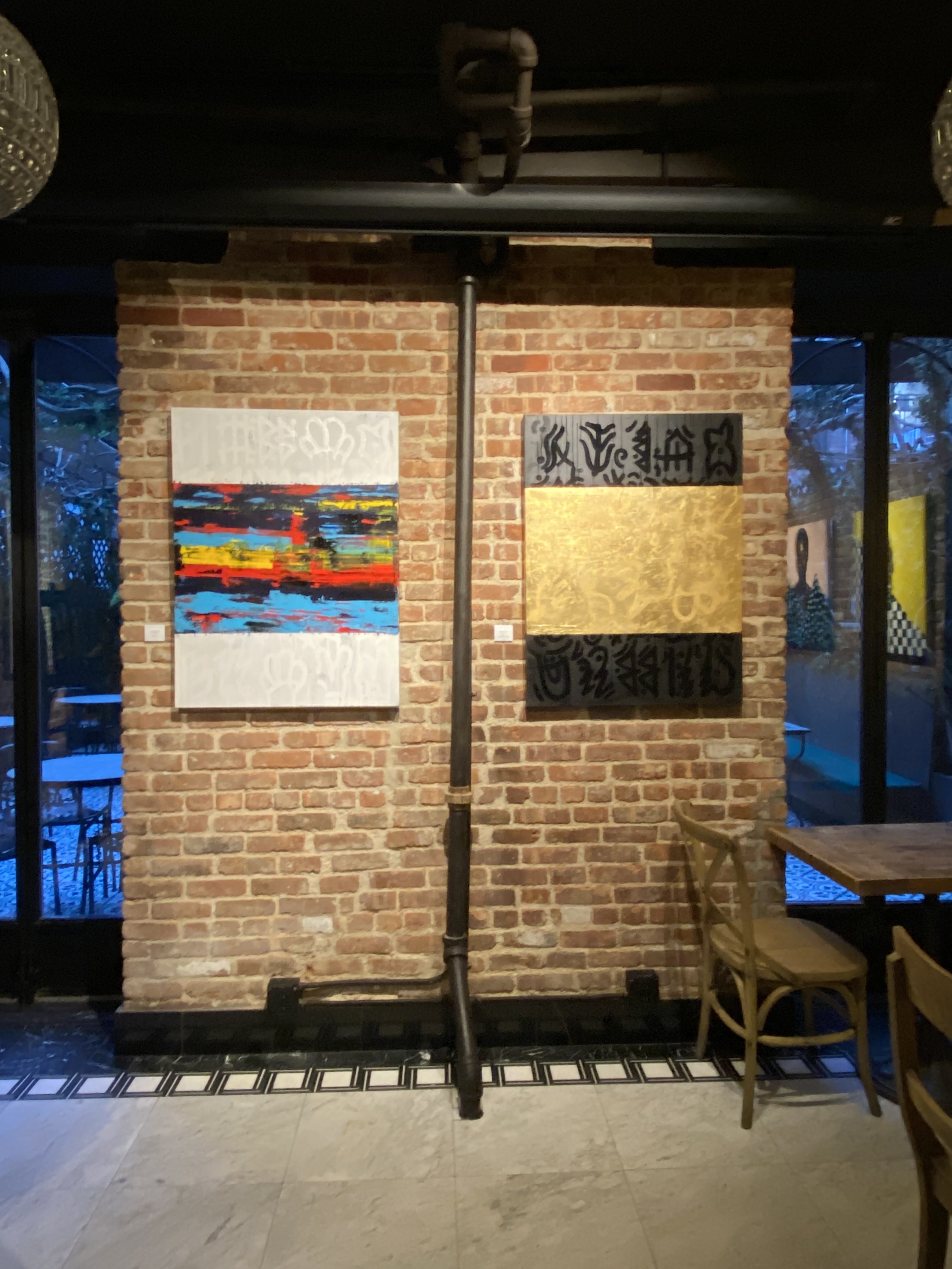 Installation view — a multicolored abstract painting beside a gold-leaf and black calligraphic work on an exposed brick wall in a dim gallery restaurant
