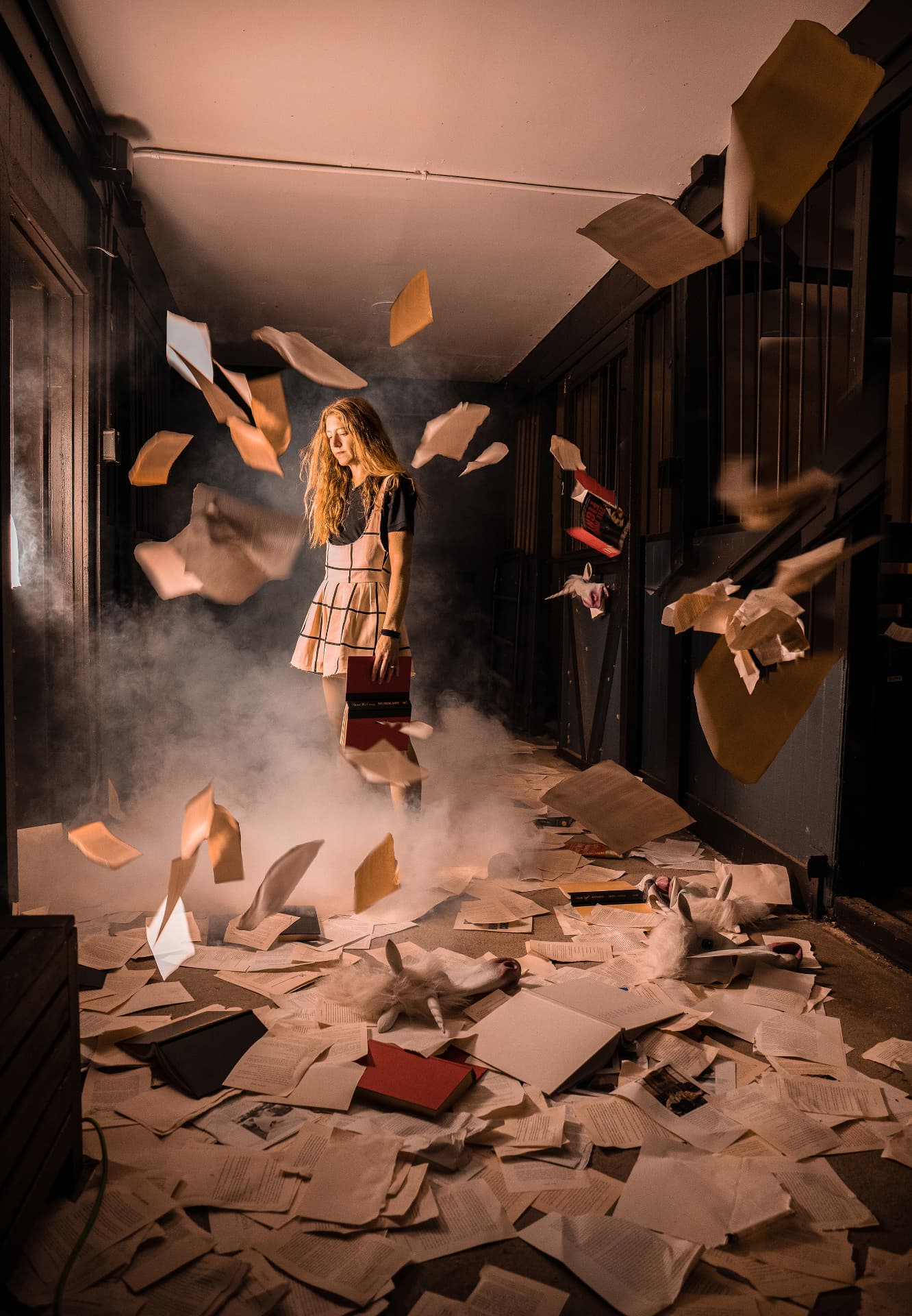 On Being — staged self-portrait of a woman in a plaid pinafore holding books amid a storm of flying papers and fog in a dark corridor