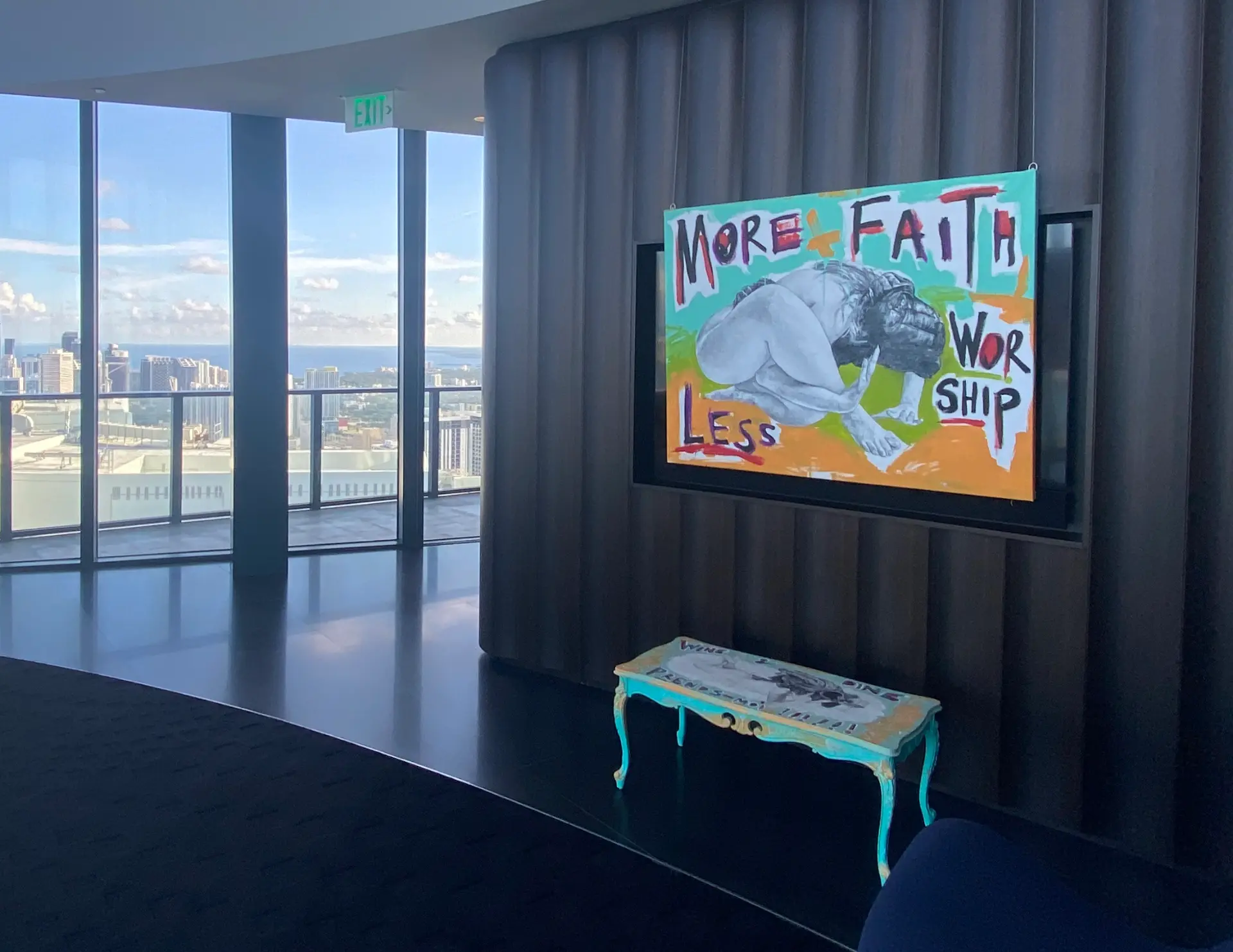 Text-based painting reading More Faith Less Worship with a reclining graphite figure, hung in a curved high-rise lounge overlooking the bay beside a hand-painted console