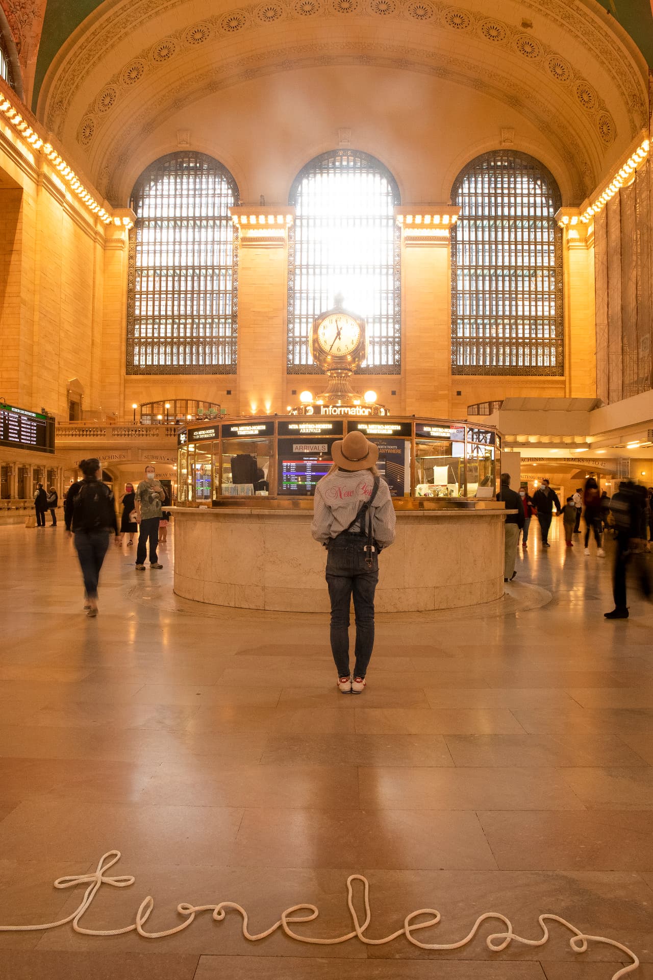 Timeless — a woman in a hat stands facing the golden information clock of Grand Central Terminal, with a rope spelled out as the word timelesss on the marble floor