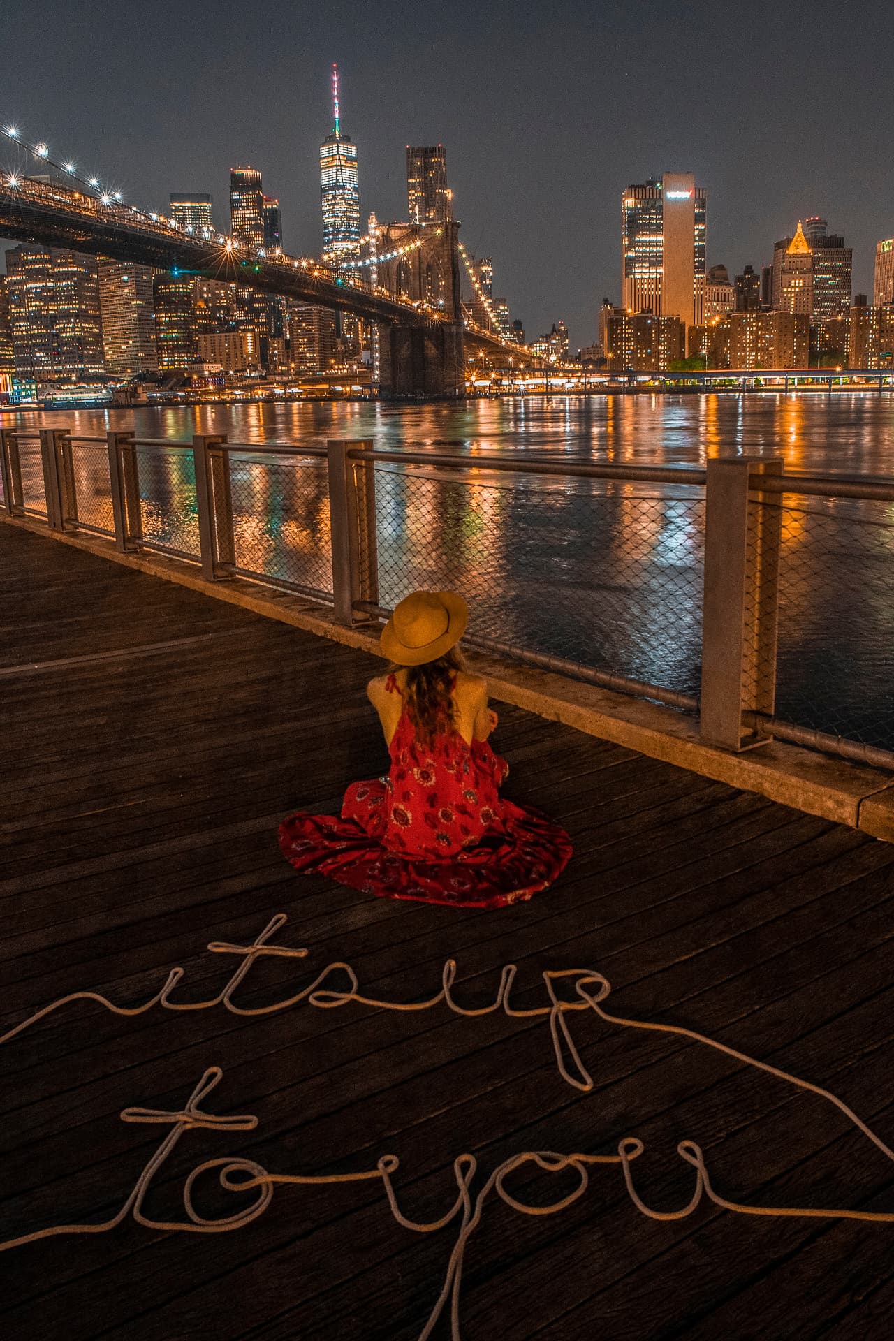 It's Up To You — a woman in a red dress sits before the illuminated Brooklyn Bridge and Manhattan skyline at night, with rope spelled out reading its up to you