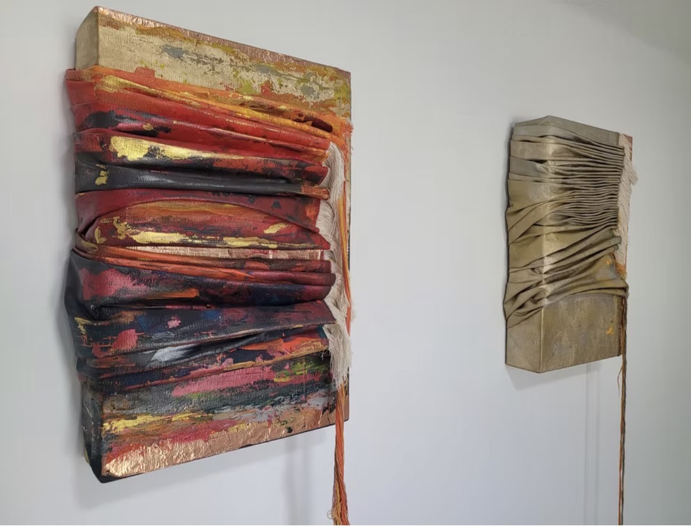 Draped painted-fabric relief with gold leaf beside a pleated golden companion work on a white gallery wall